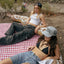 Two women sitting on a checkered blanket outdoors, one wearing a white crop top and black pants, the other in a black bikini top and denim shorts.